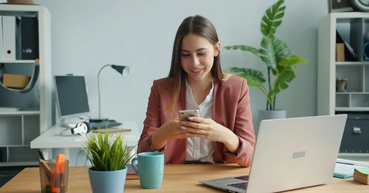 Mujer de negocios sonríe al leer un mensaje en su teléfono, con la notebook abierta en el escritorio
